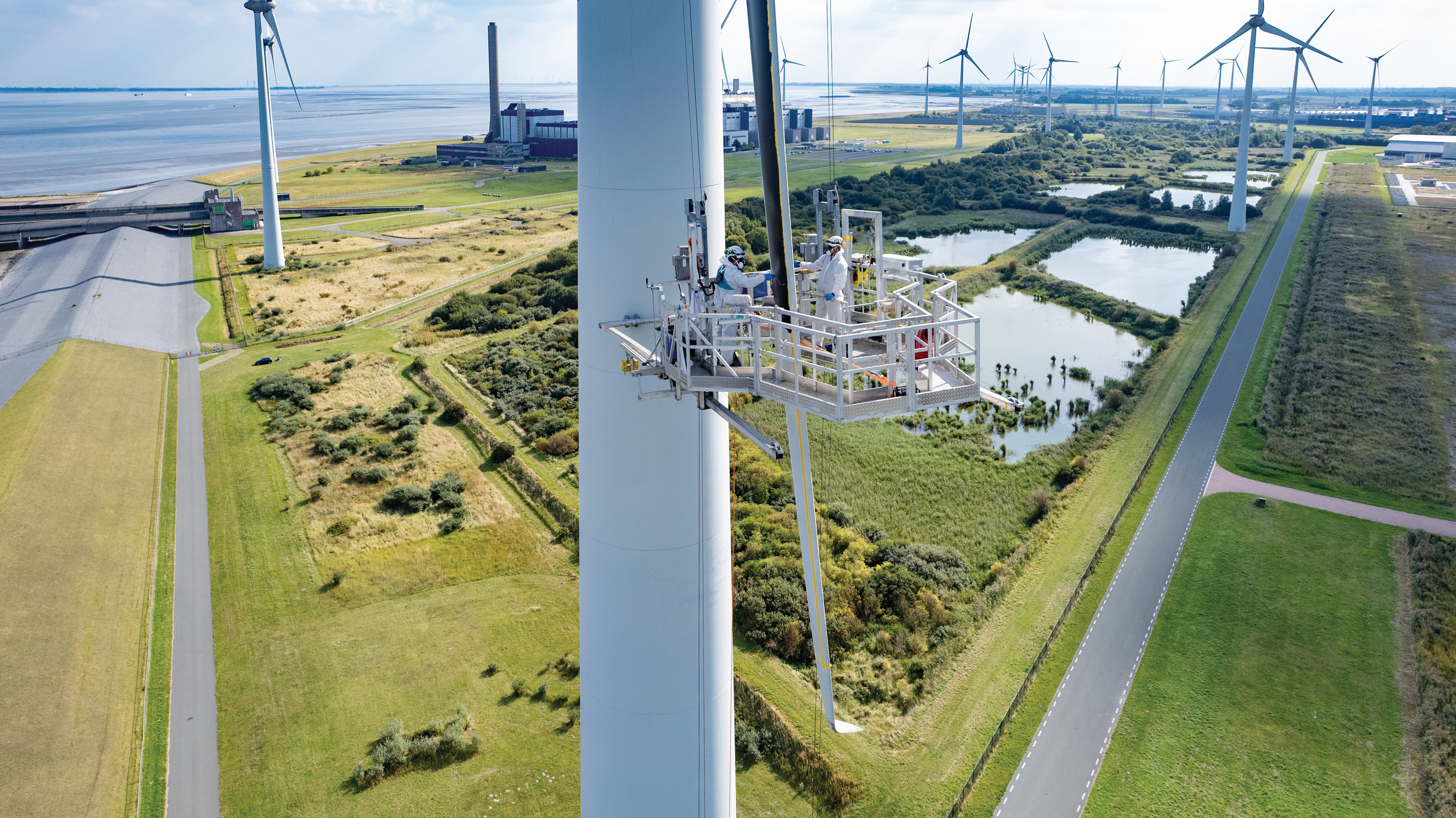 A wind turbine being serviced from a platform. In the background, there are additional turbines and green landscapes.