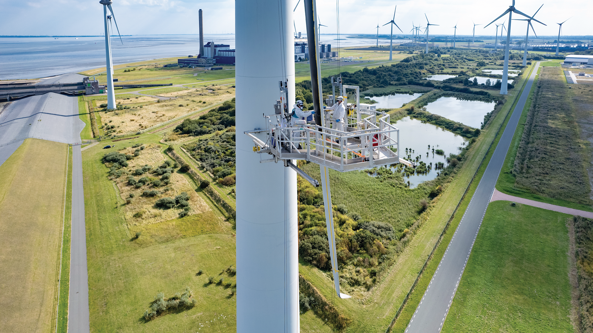 A wind turbine being serviced from a platform. In the background, there are additional turbines and green landscapes.