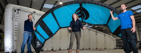 Drei Personen im Innenraum halten einen Kitesurf-Schirm. Der Schirm ist schwarz und türkis mit einem auffälligen Design.
