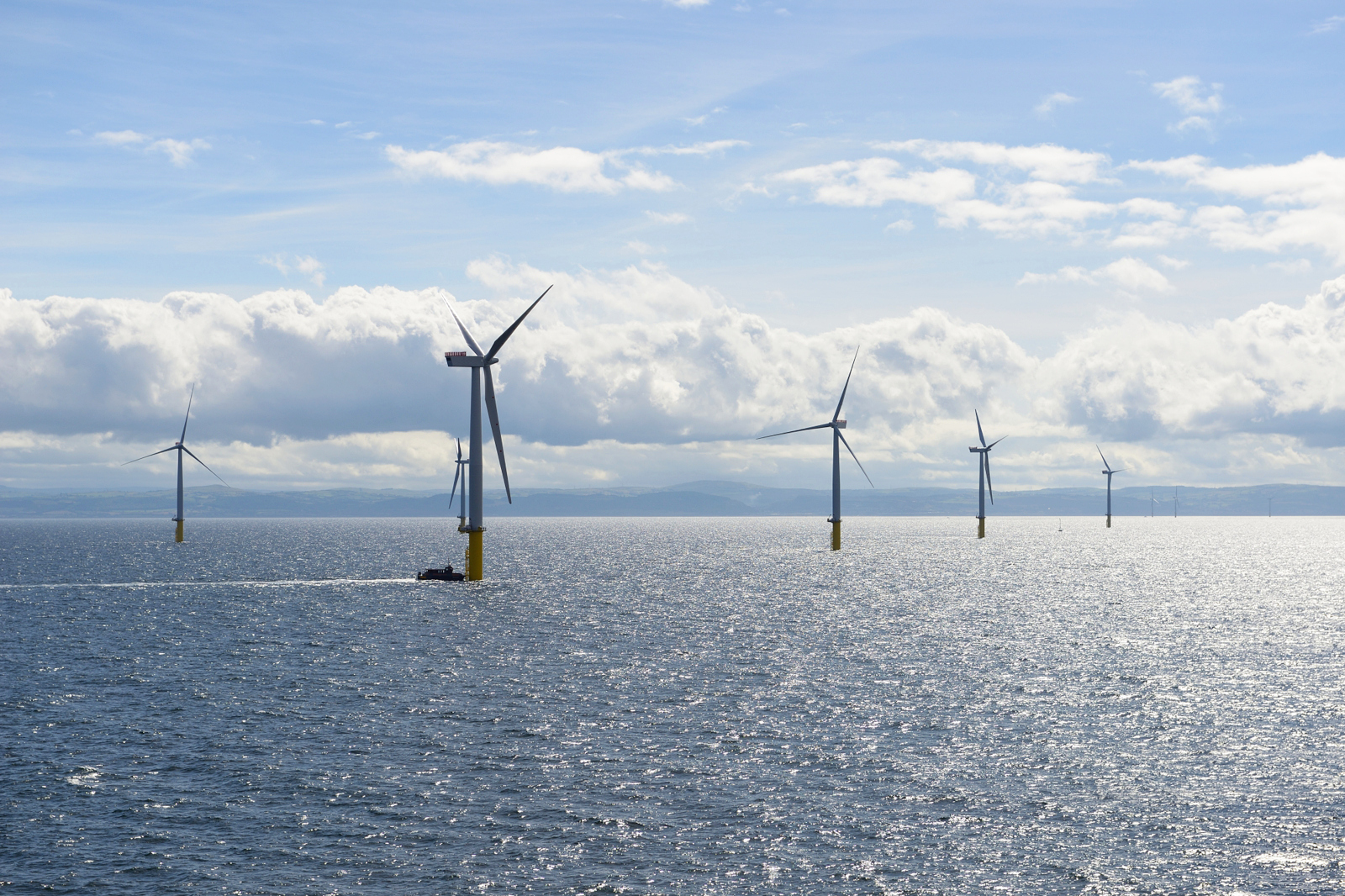 Weiße Offshore-Windturbinen mit gelben Sockeln stehen im sonnenfunkelnden blau-grauen Meer unter einem teils bewölkten Himmel.