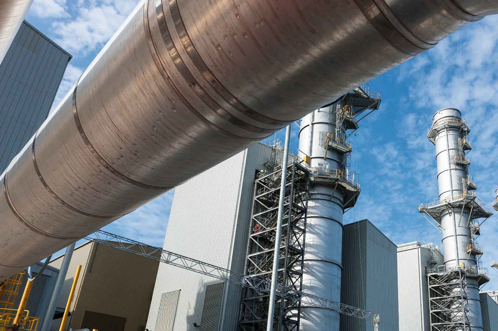 Eine Nahaufnahme von großen Stahlrohren an einem Industriestandort mit sichtbaren Rauchschornsteinen unter einem blauen Himmel.