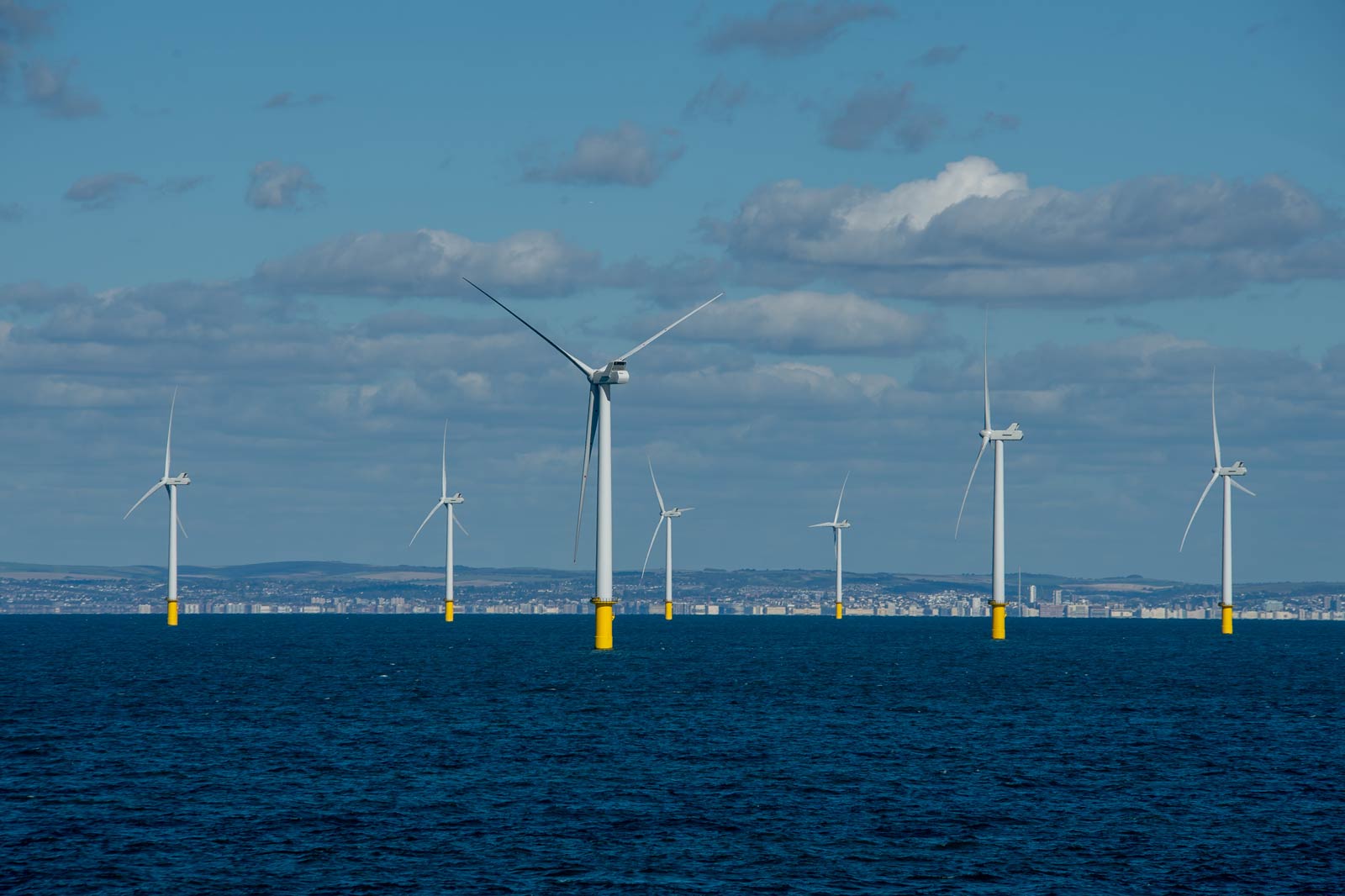 Eine Reihe von Windturbinen auf dem Wasser mit gelben Sockeln vor einem klaren Himmel und einer Stadt im Hintergrund.