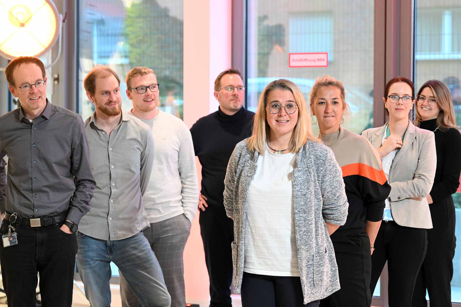 Eine Gruppe von Menschen steht in einem hellen Raum mit großen Fenstern. Die Personen tragen legere Kleidung.