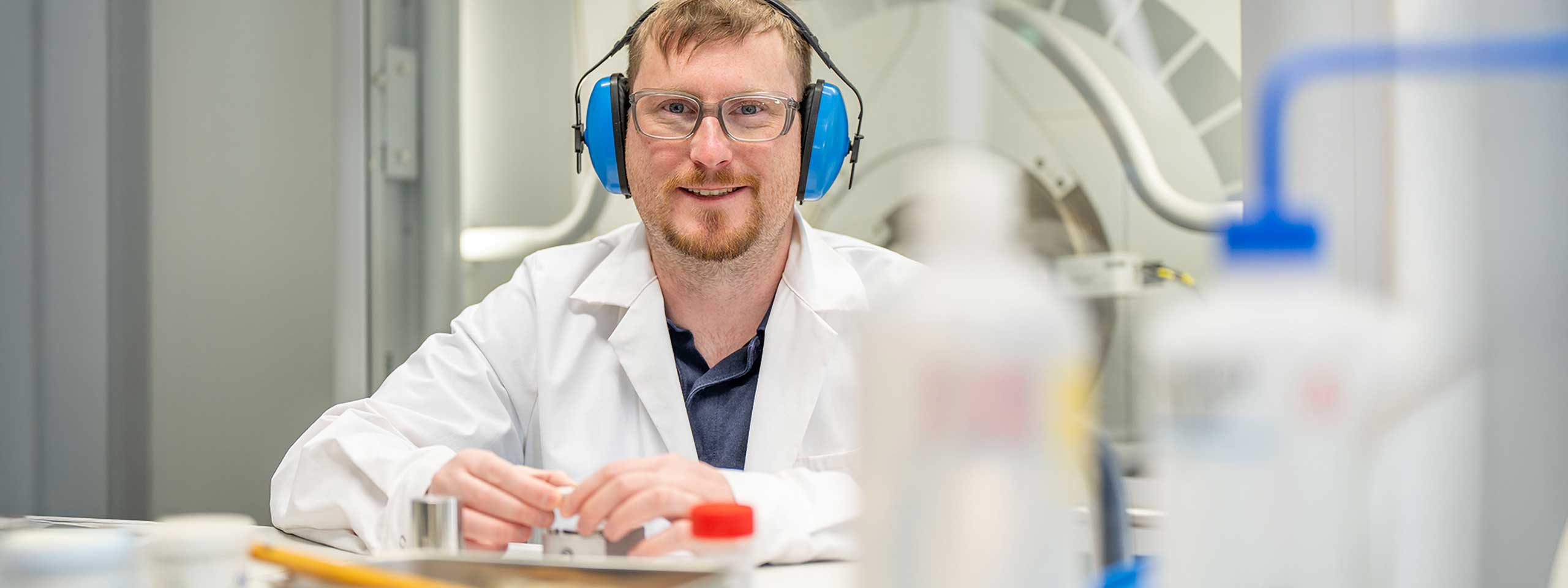 Person im weißen Laborkittel mit blauem Gehörschutz und Händen auf dem Tisch.