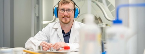 Person im weißen Laborkittel mit blauem Gehörschutz und Händen auf dem Tisch.