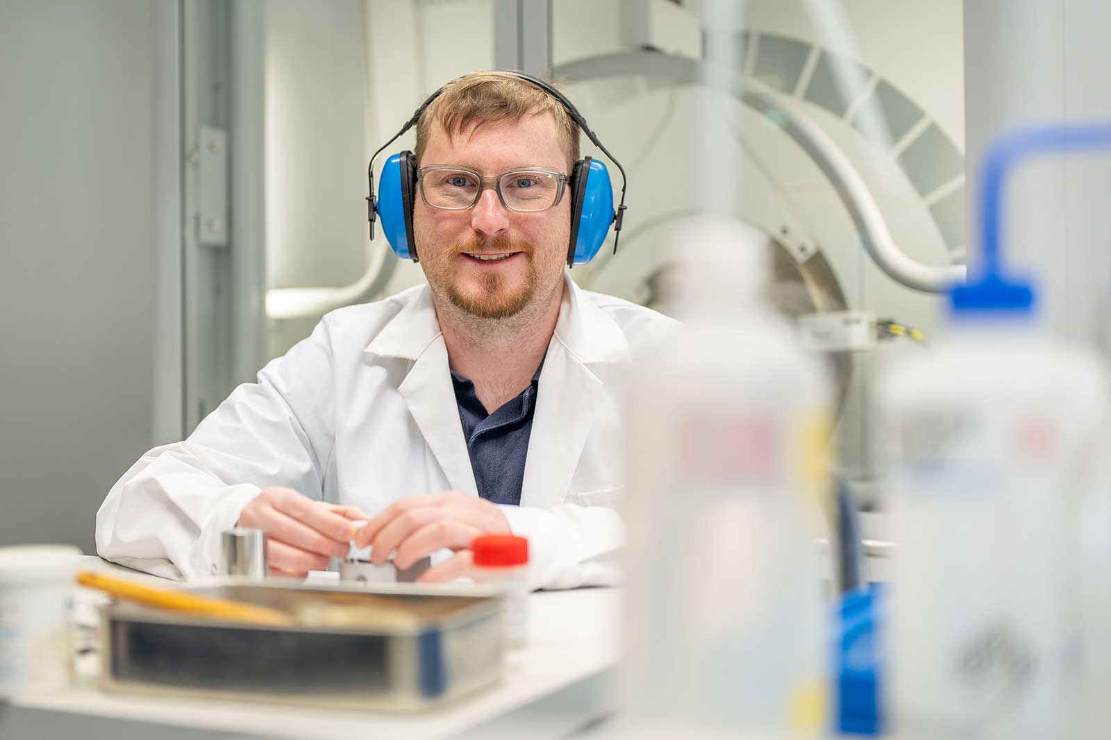 Person im weißen Laborkittel mit blauem Gehörschutz und Händen auf dem Tisch.