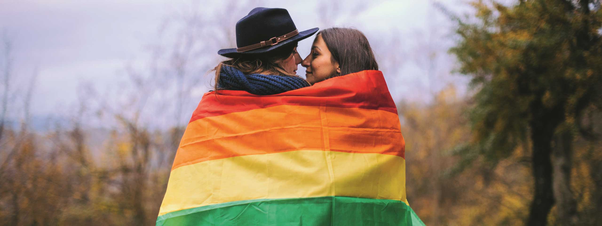 Ein Paar steht in einer Umarmung, eingehüllt in eine Regenbogenflagge, vor einer herbstlichen Landschaft.