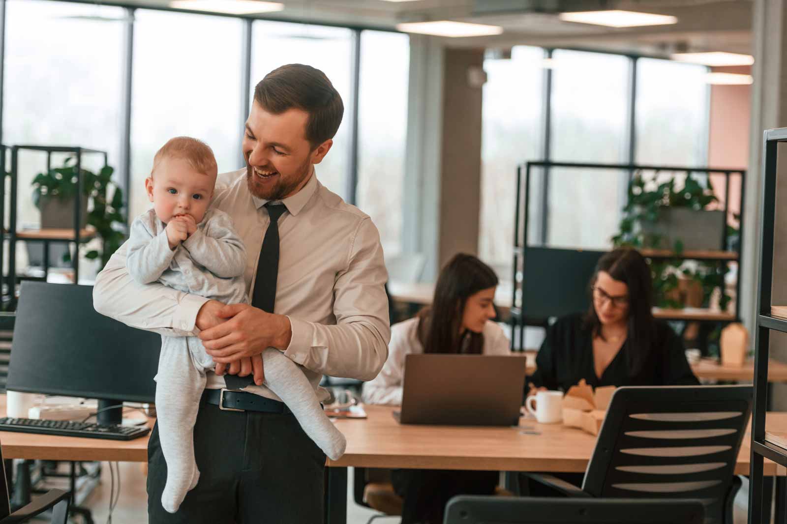 Ein Mann im Hemd und mit Krawatte hält ein Baby. Im Hintergrund arbeiten zwei Frauen an Laptops in einem modernen Büro.