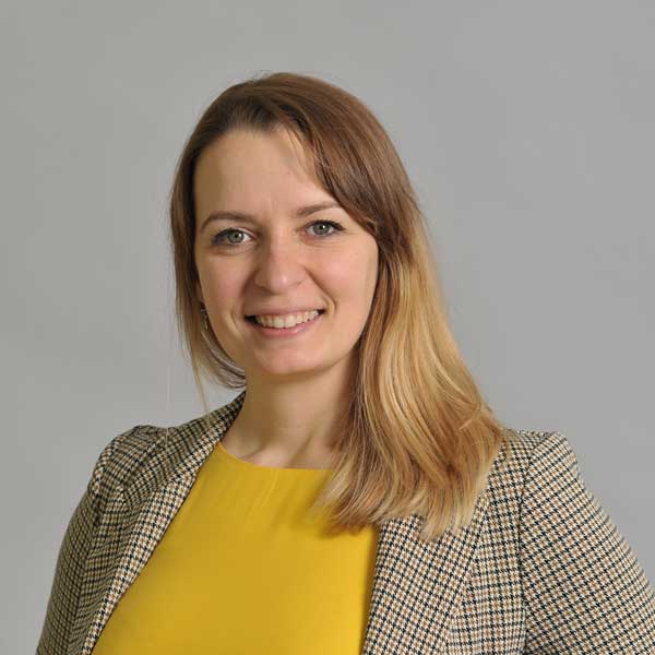 Eine Person trägt ein kariertes Blazer und ein gelbes Oberteil vor einem grauen Hintergrund. Portrtait von Ivana Rosenboom.
