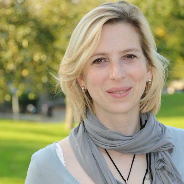 Eine Person mit blonden, mittellangen Haaren trägt ein graues Tuch und steht in einer grünen Umgebung mit Bäumen. Portrtait von Melanie Lober.