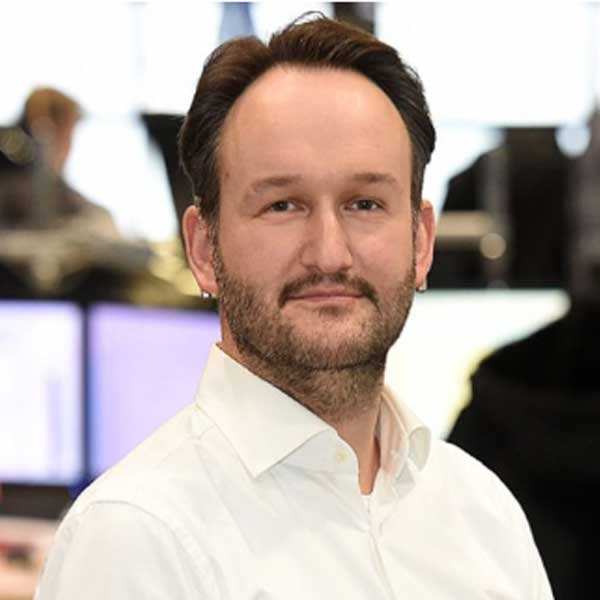 Ein Mann in einem weißen Hemd steht in einem Büro mit Computerbildschirmen im Hintergrund. Portrait von Matthias Schröder.