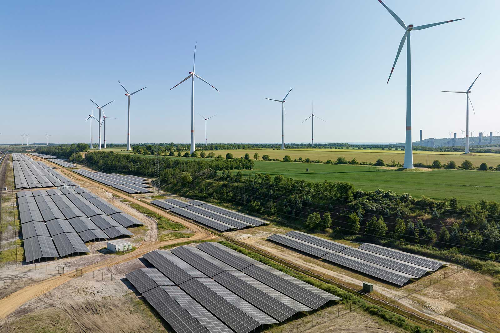 Eine Landschaft mit Solaranlagen im Vordergrund und Windkraftanlagen im Hintergrund unter klarem Himmel.