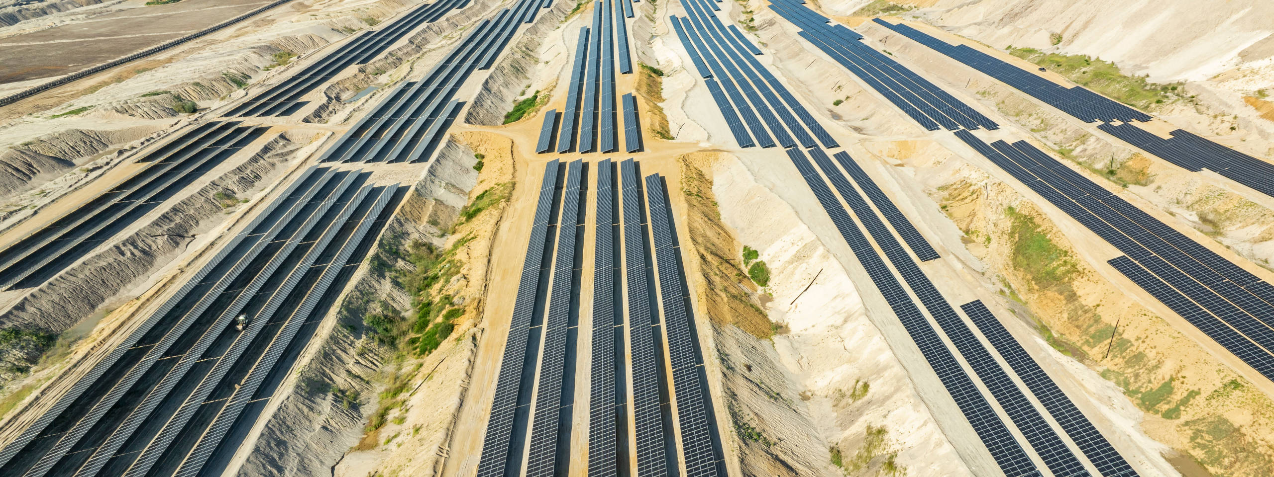 Ein Luftbild einer Solaranlage mit zahlreichen Solarpanels, die in schmalen Reihen in einem sandigen Gelände angeordnet sind.