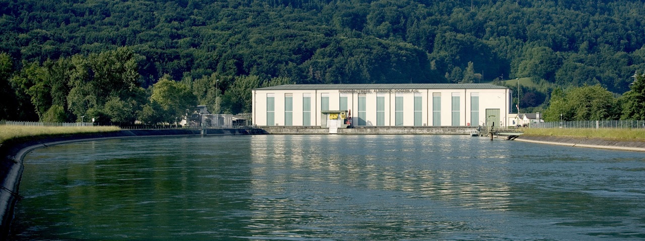 Ein modernes Gebäude an einem Gewässer, umgeben von Bäumen und Hügeln, reflektiert im ruhigen Wasser.