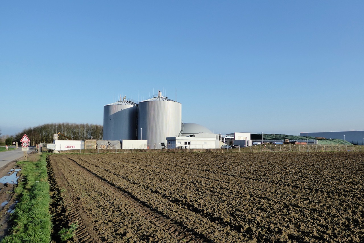 Eine Biogasanlage mit großen Tanks steht neben einem frisch bearbeiteten Feld. Der Himmel ist klar und blau.