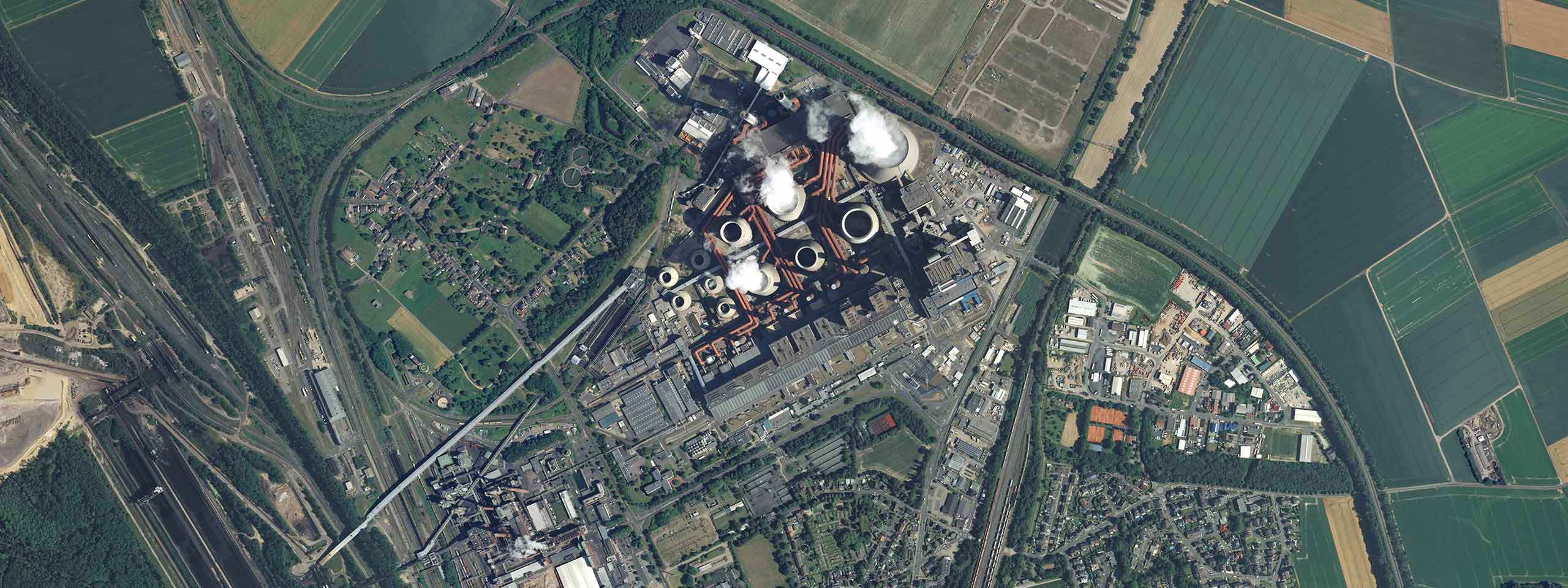 Luftbild einer Industrieanlage mit Wassertürmen, dampfenden Schloten und umliegenden Feldern sowie Wohngebieten.