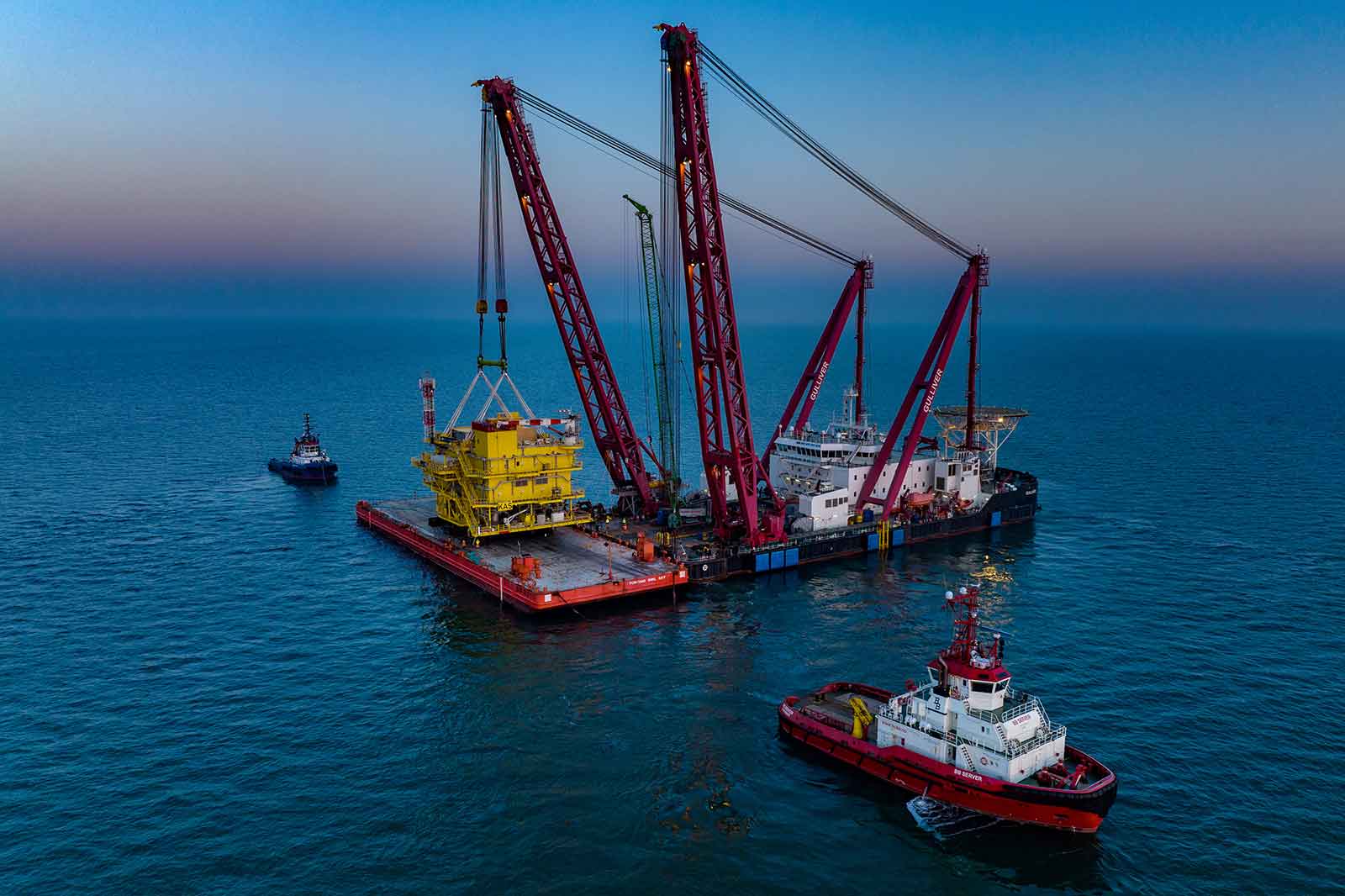 Ein beeindruckendes Offshore-Schiff hebt eine gelbe Plattform aus dem Wasser. Zwei Schlepper sind in der Nähe.