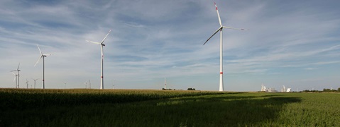 Eine Landschaft mit mehreren Windkraftanlagen und einer Weizenfeld im Vordergrund. Im Hintergrund sind Fabriken sichtbar.