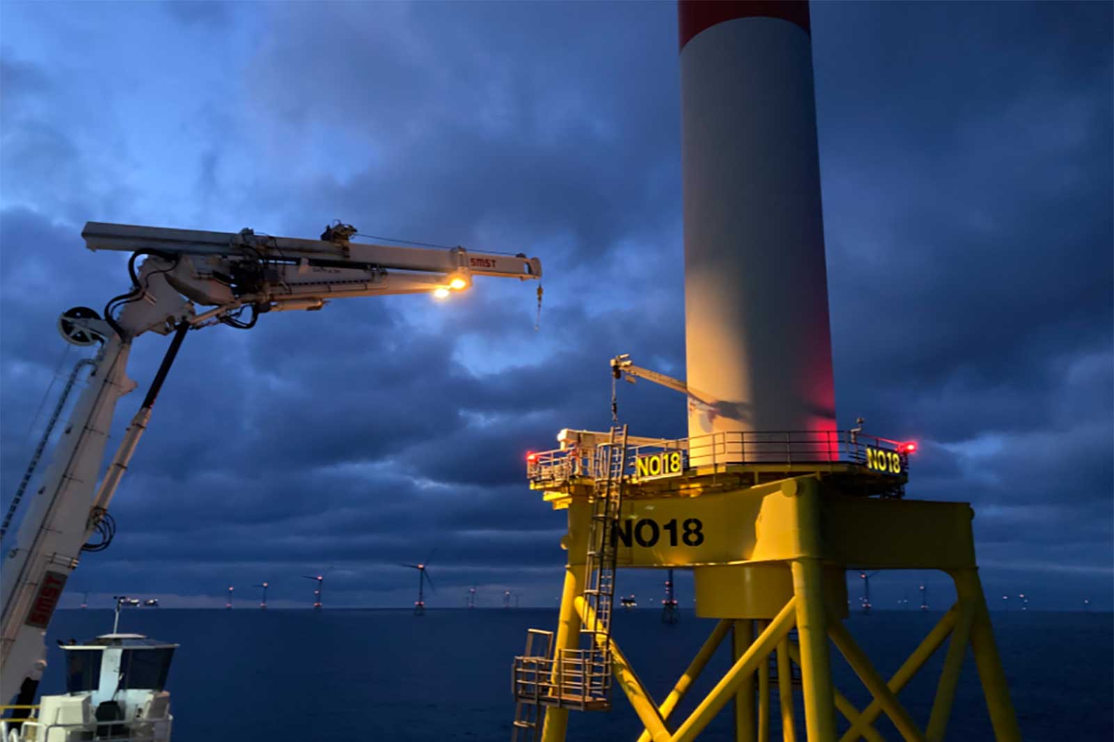 Ein Ausblick auf eine Offshore-Windkraftanlage bei Dämmerung, mit einem Kranarm und deutlich sichtbaren Windturbinen im Hintergrund.