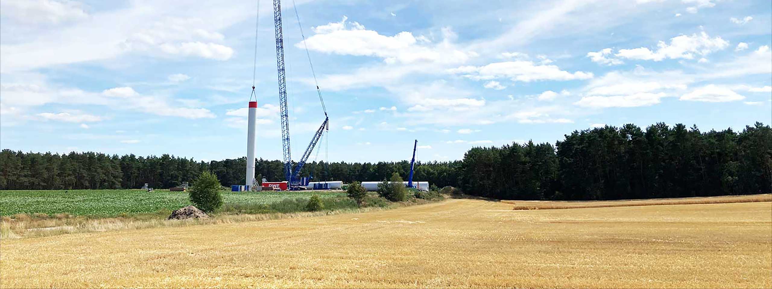 Ein Windrad wird auf einem Feld mit einem Kran errichtet. Im Hintergrund sind Wälder und blauer Himmel zu sehen.