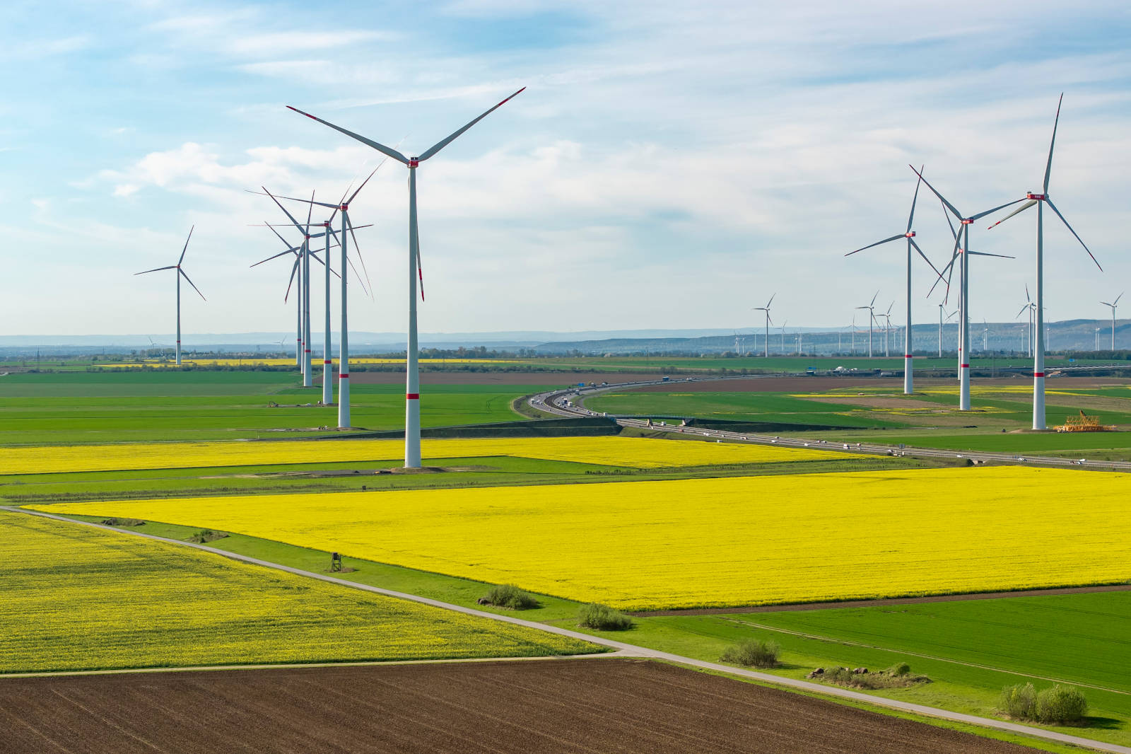 Mehrere Windräder stehen auf grünen und leuchtend gelben Rapsfeldern neben einer Landstrasse mit fernen Fahrzeugen unter leicht bewölktem Himmel.