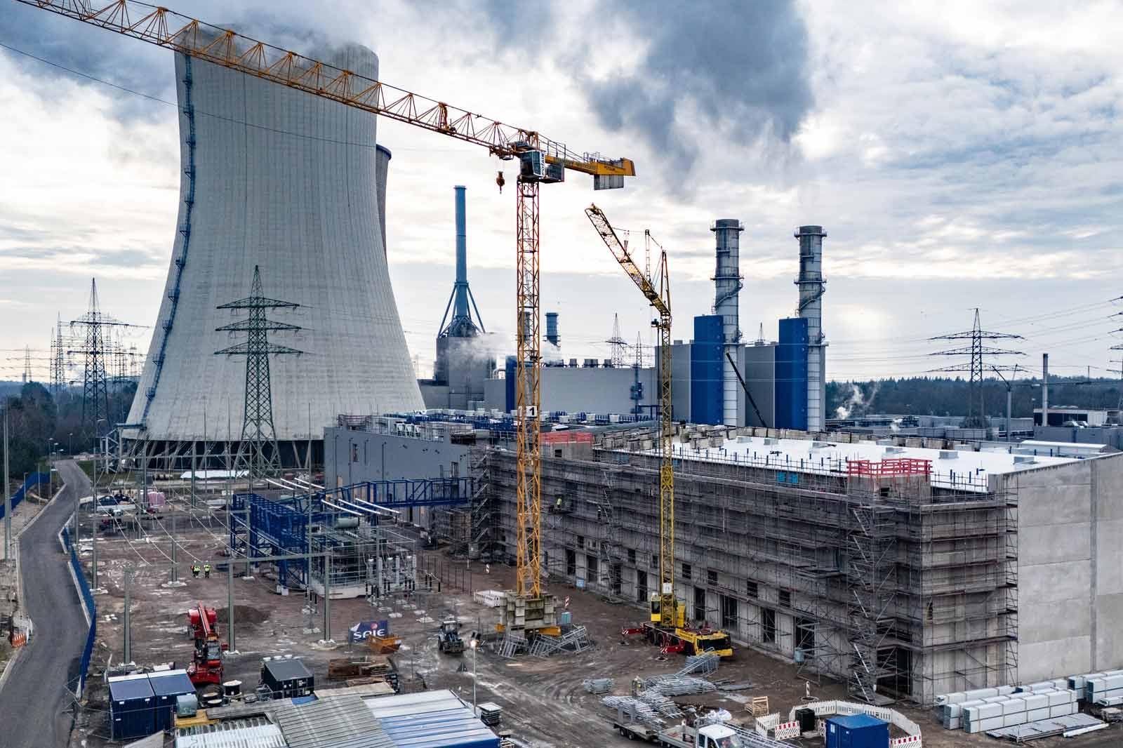 Eine Baustelle mit einem Kühlturm, Kränen und Industriebauten. Rauch und Stromleitungen sind im Hintergrund sichtbar.