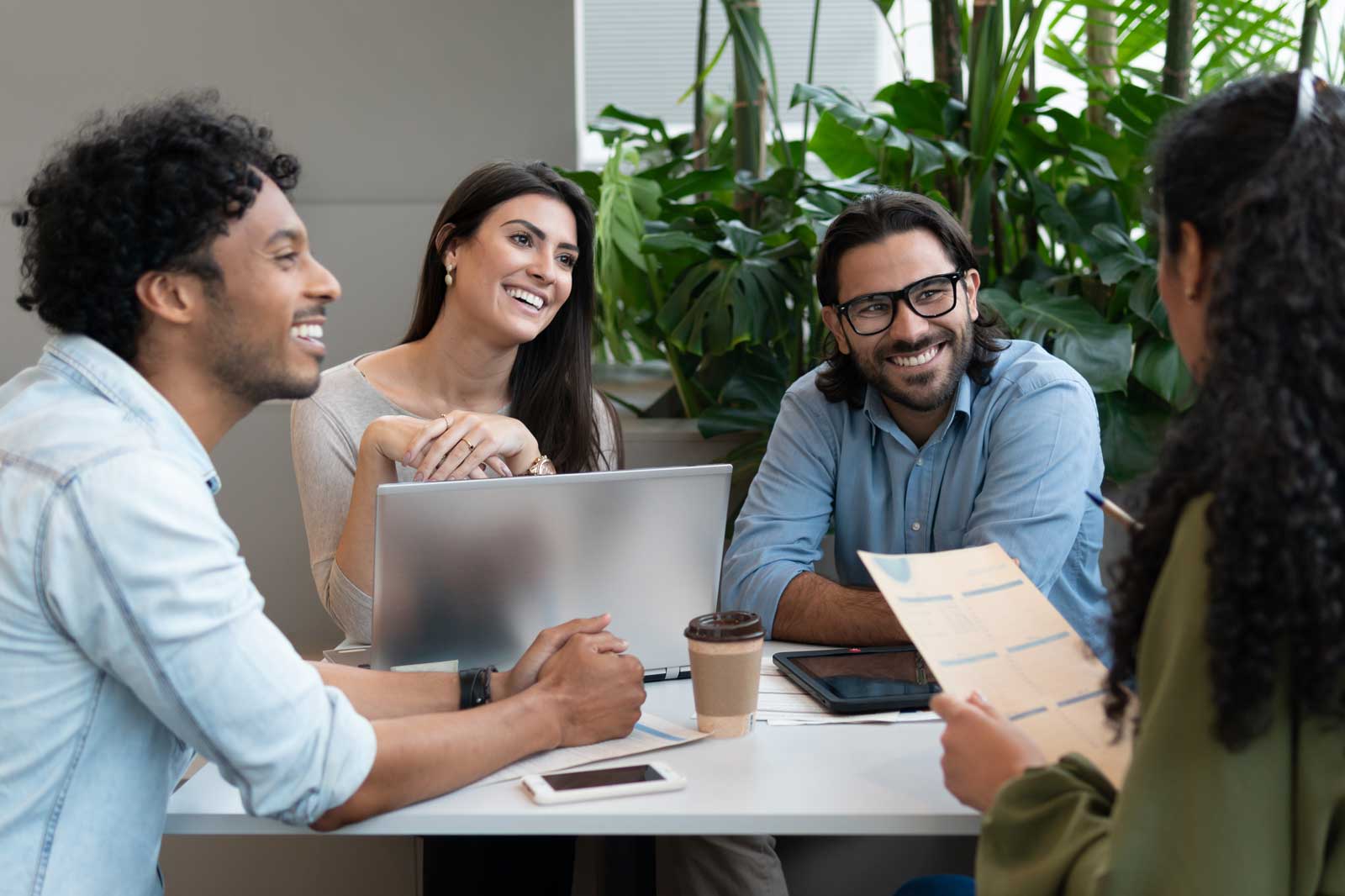 A diverse group of four individuals engaged in a collaborative meeting around a table with plants in the background.