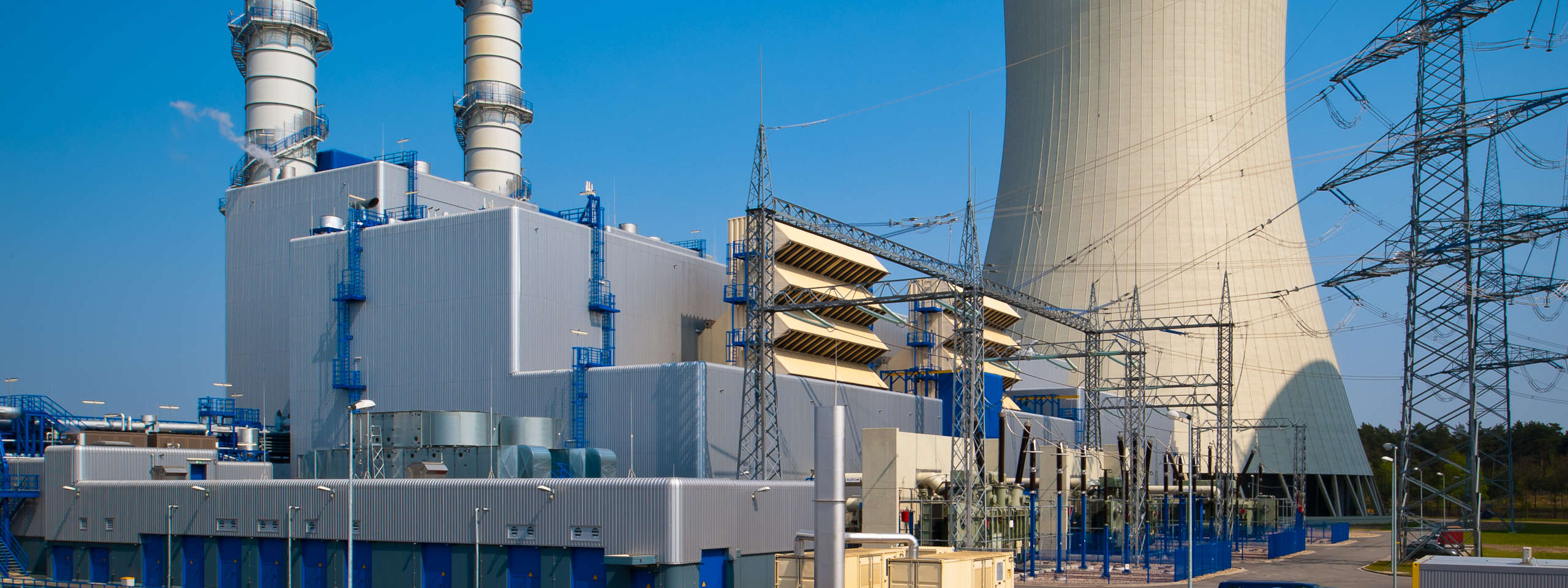 An industrial complex featuring a cooling tower, smokestack, and electrical installations under a clear blue sky.
