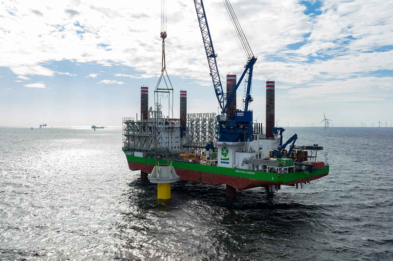 An offshore ship with a large crane lifts a yellow structure from the water, surrounded by wind turbines in the background.