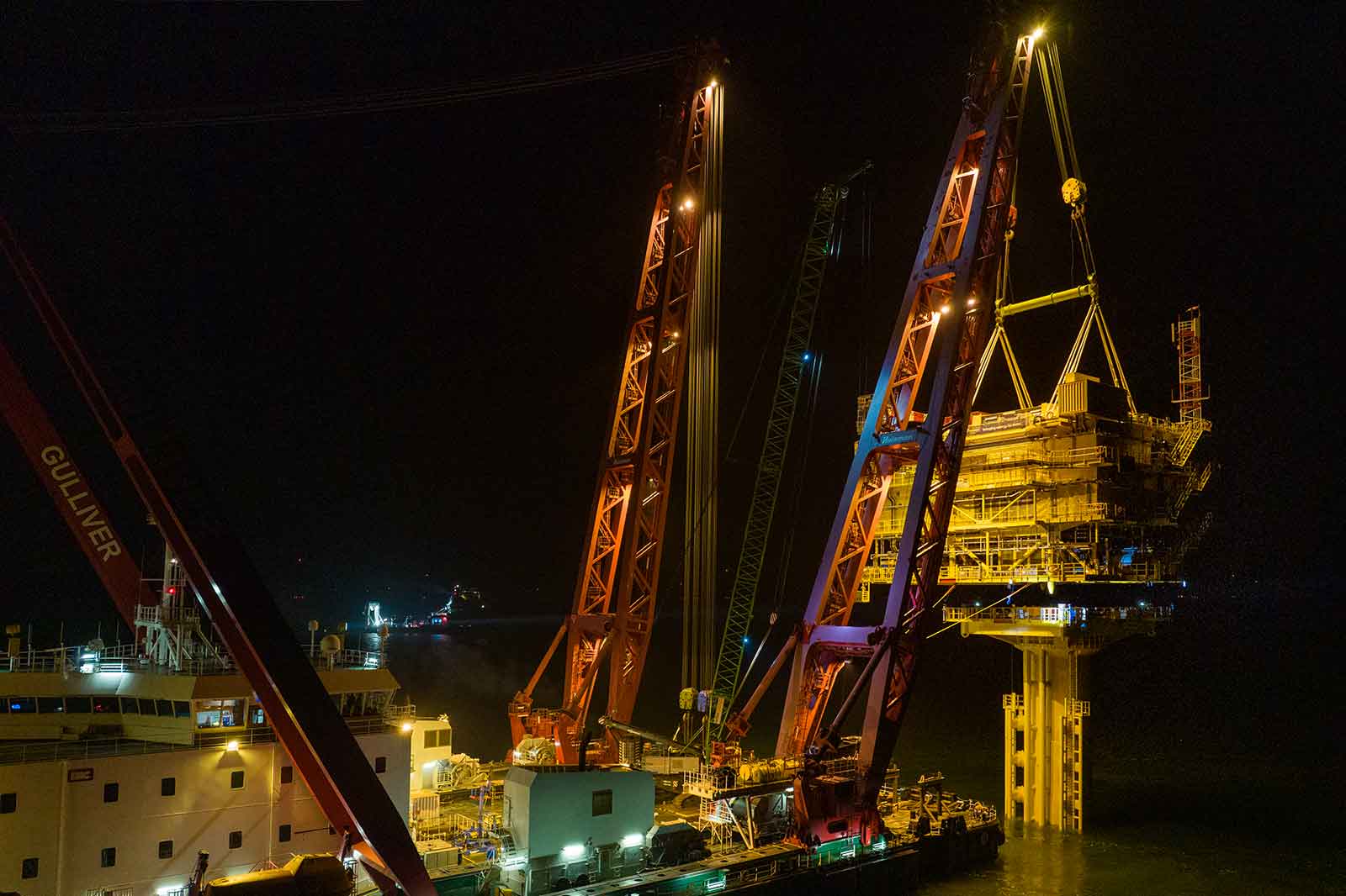 An offshore vessel lifts a platform at night with orange cranes. The water reflects the lights.