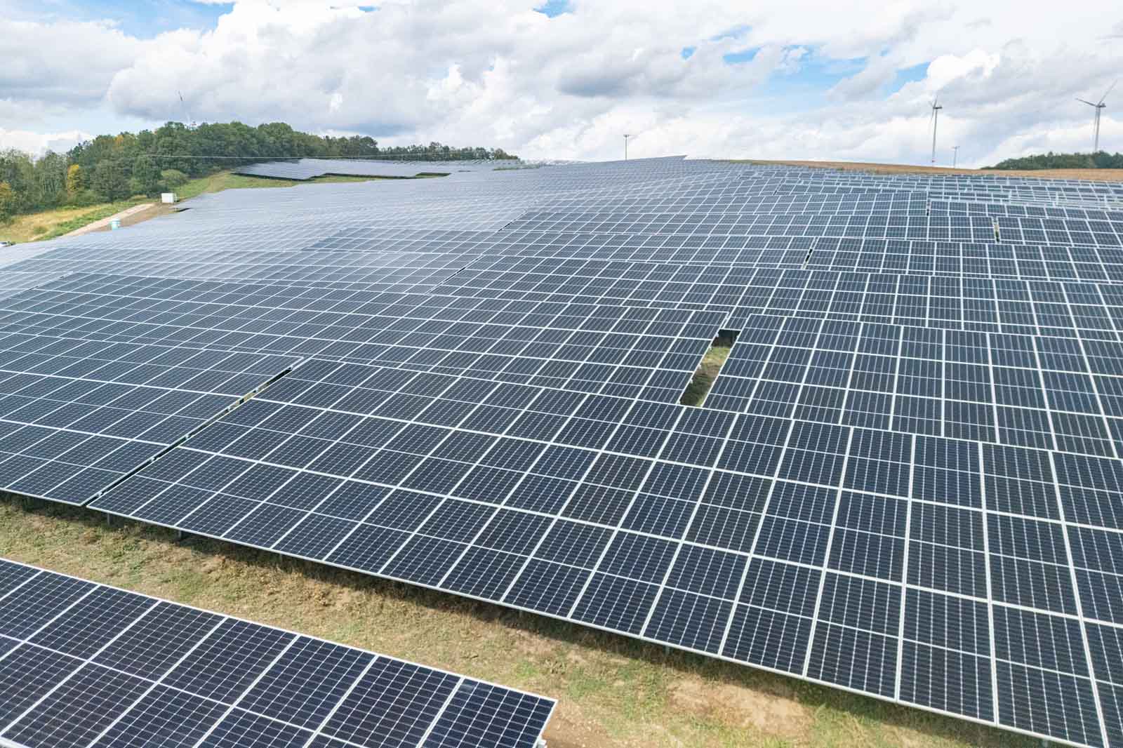 Ein weitläufiges Solarpanelfeld unter einem teilweise bewölkten Himmel, mit grünen Hügeln im Hintergrund.