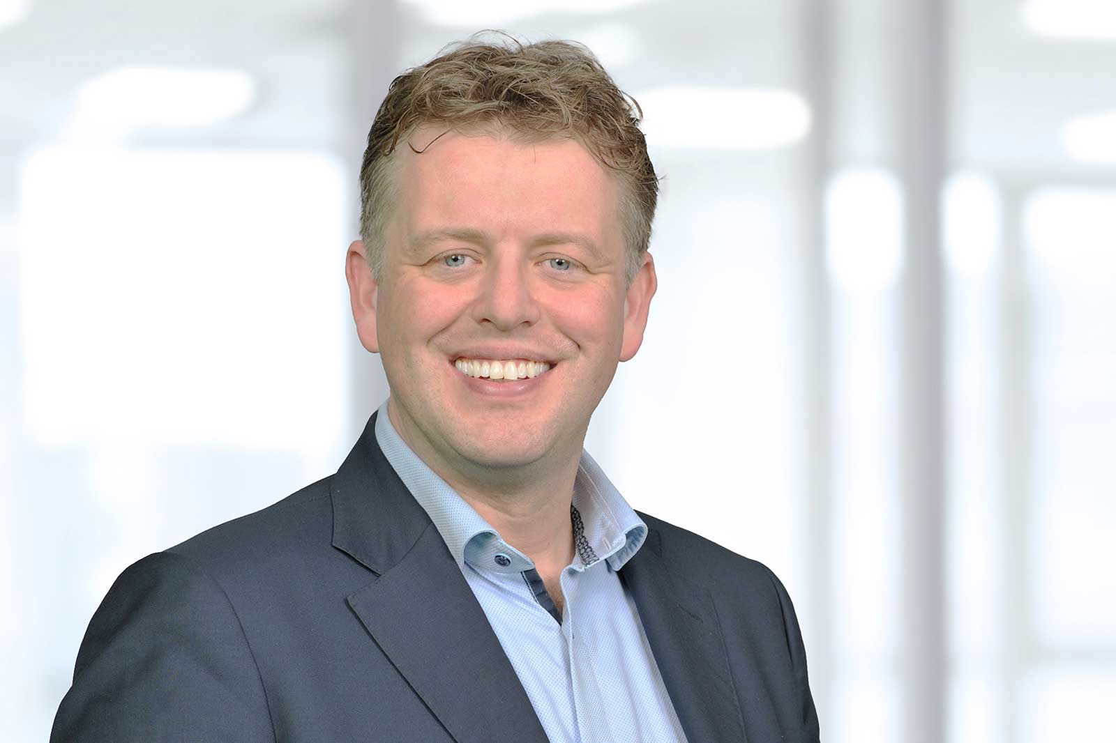 A man in a suit with a blue shirt stands against a bright, blurred background.