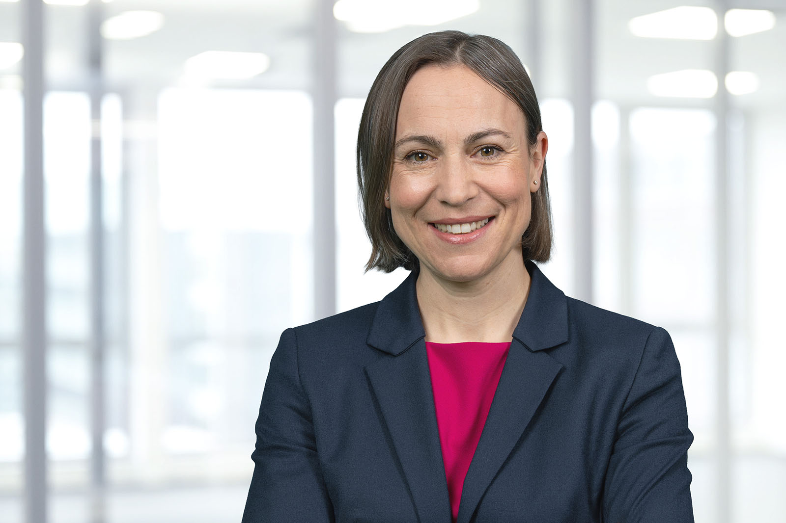 Eine Frau mit kurzem dunklem Haar in einem marineblauen Blazer und einer magentafarbenen Bluse steht mit verschränkten Armen in einem hellen Büro mit großen Fenstern. Portrait von Sandra Dettmer.