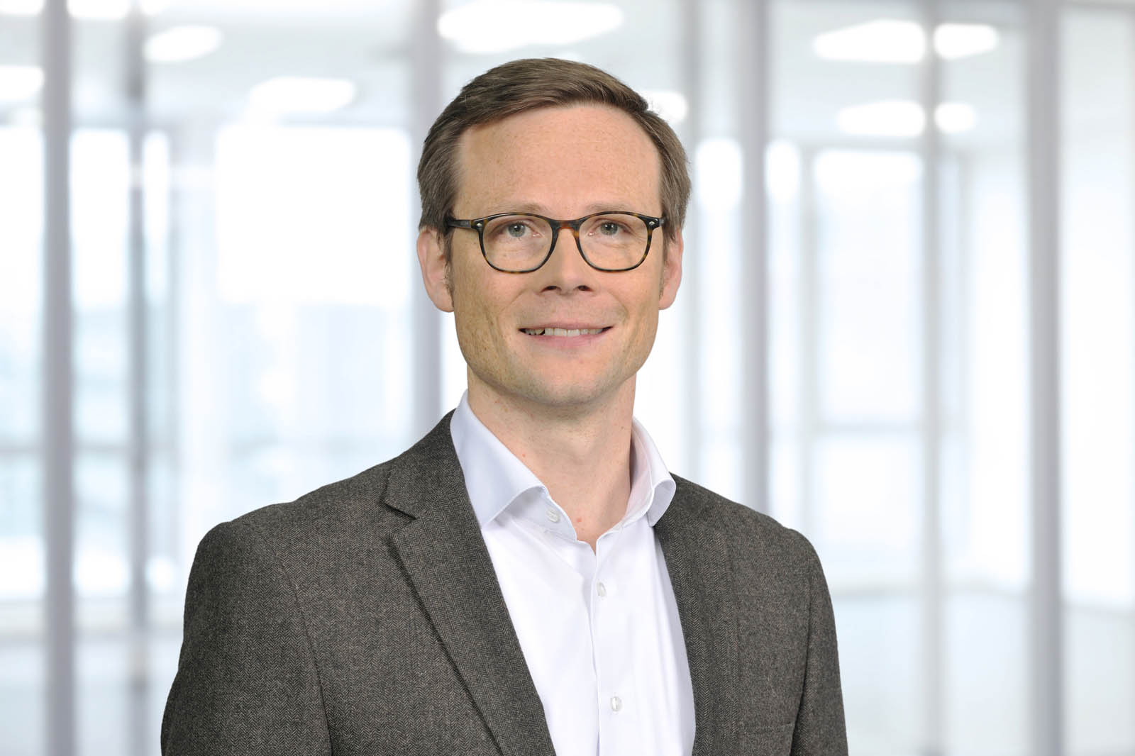 Ein Mann in einem grauen Blazer und einem weißen Hemd steht vor einem modernen, lichtdurchfluteten Hintergrund.