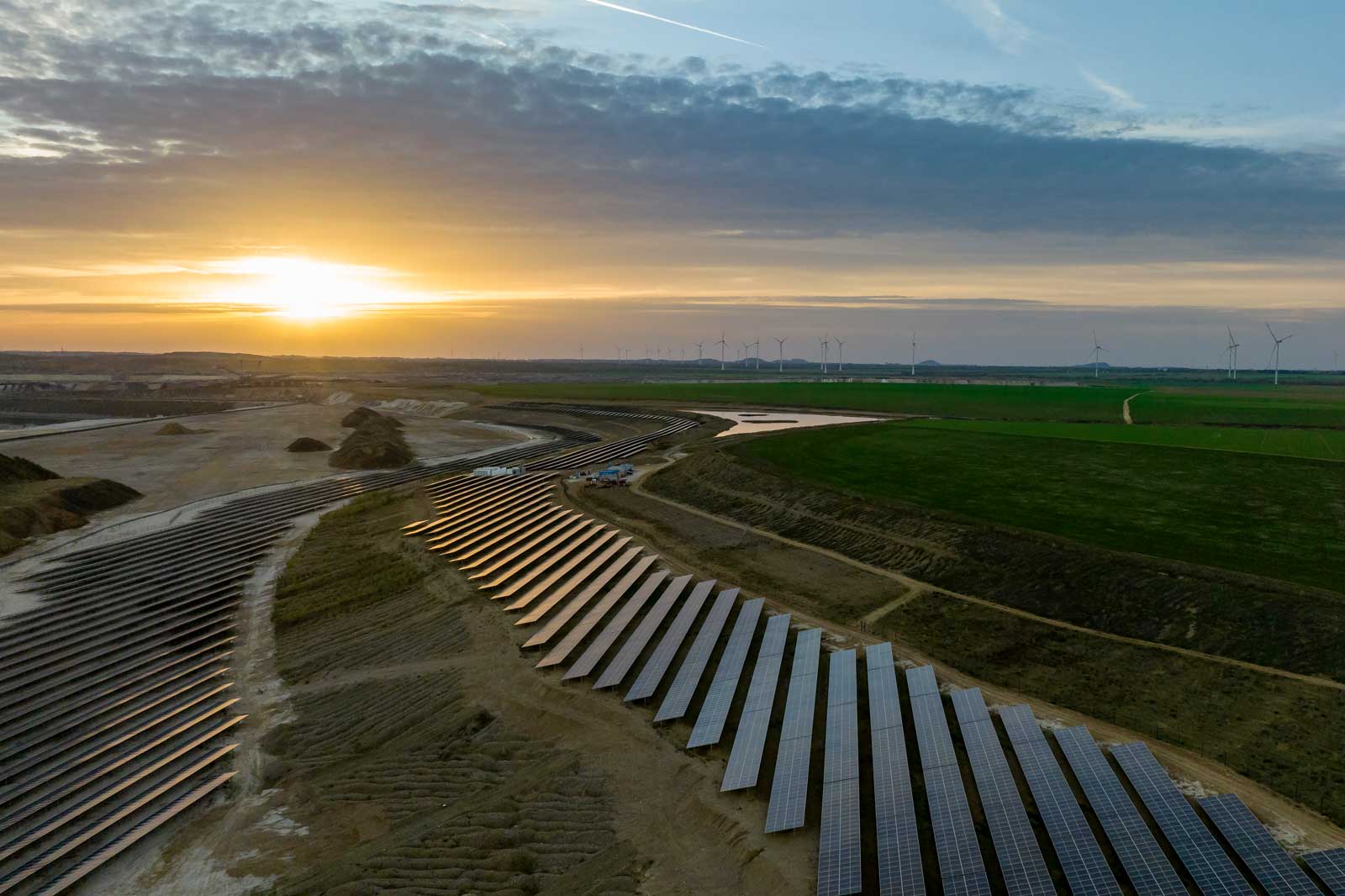 Weitwinkelaufnahme eines Solarkraftwerks bei Sonnenuntergang mit Windkraftanlagen im Hintergrund.