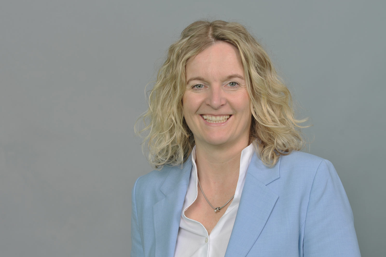 Eine Person mit lockigem, blondem Haar trägt einen hellblauen Anzug und ein weißes Hemd vor einem grauen Hintergrund. Portrait von Anja Dubbert.