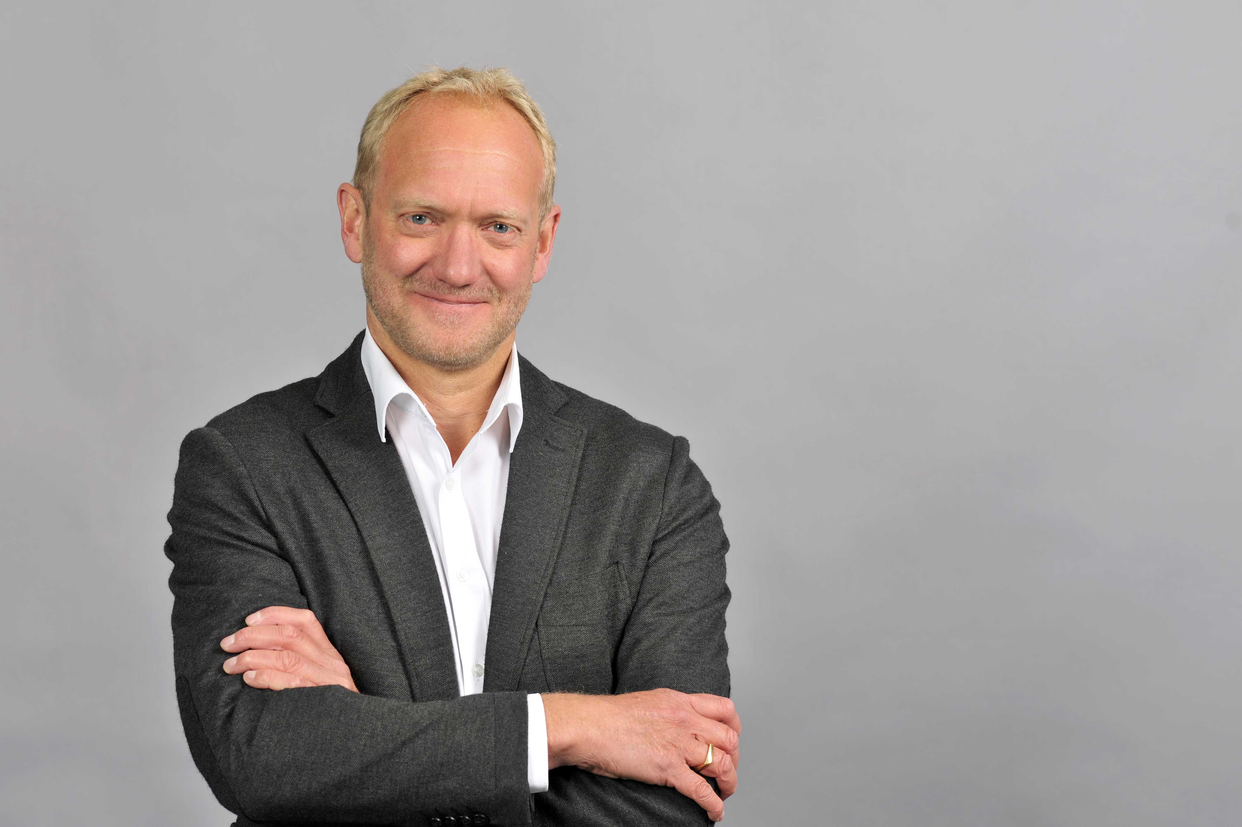 Eine Person mit einem grauen Anzug und weißem Hemd steht mit verschränkten Armen vor einem grauen Hintergrund. Portrait von Ansgar Vierhaus.