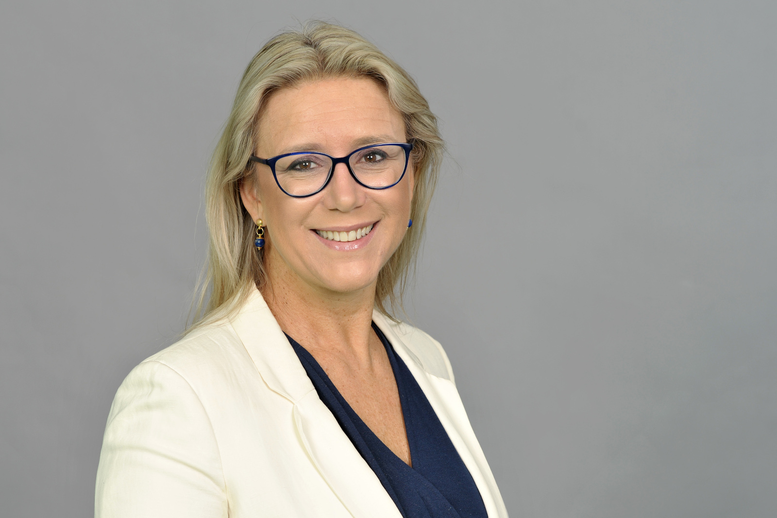 Eine Frau mit langen, blonden Haaren trägt einen Blazer über einem marineblauen Oberteil vor einem grauen Hintergrund. Portrait von Federica Maranca.