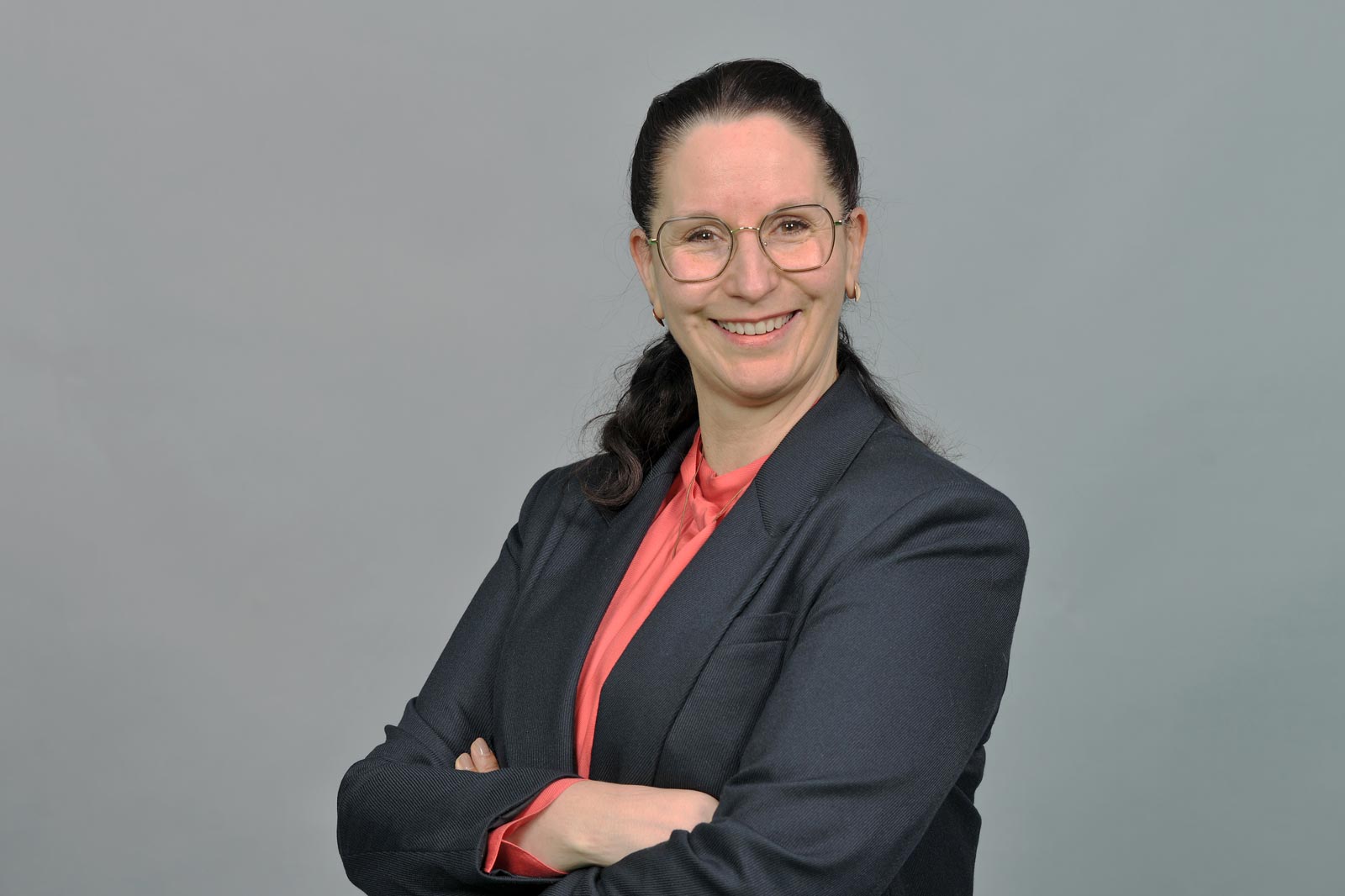 Eine Frau mit dunklem Haar trägt einen schwarzen Blazer und ein rotes Oberteil. Sie hat ihre Arme verschränkt. Portrait von Melanie Kleemann.