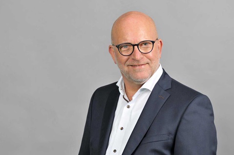 Ein Mann in einem formellen Anzug steht vor einem grauen Hintergrund, seine Gesichtszüge sind unkenntlich gemacht. Portrait von Michael Schütz.