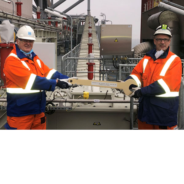 Zwei Arbeiter in schützender Kleidung halten einen großen Schraubenschlüssel auf einer industriellen Anlage.