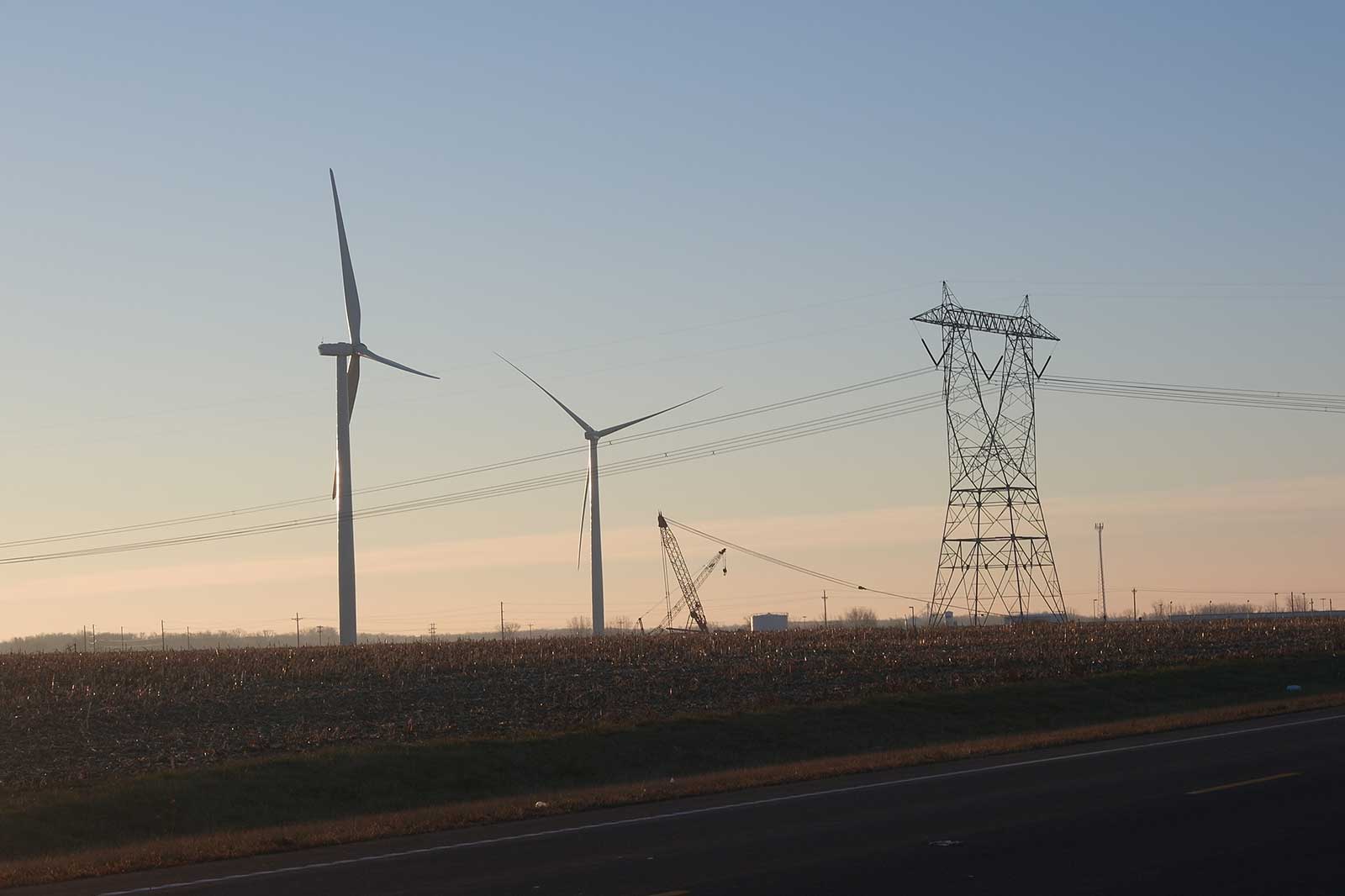Windkraftanlagen und Hochspannungsmasten in einem ländlichen Umfeld bei Sonnenaufgang, mit einer Straßenansicht im Vordergrund.