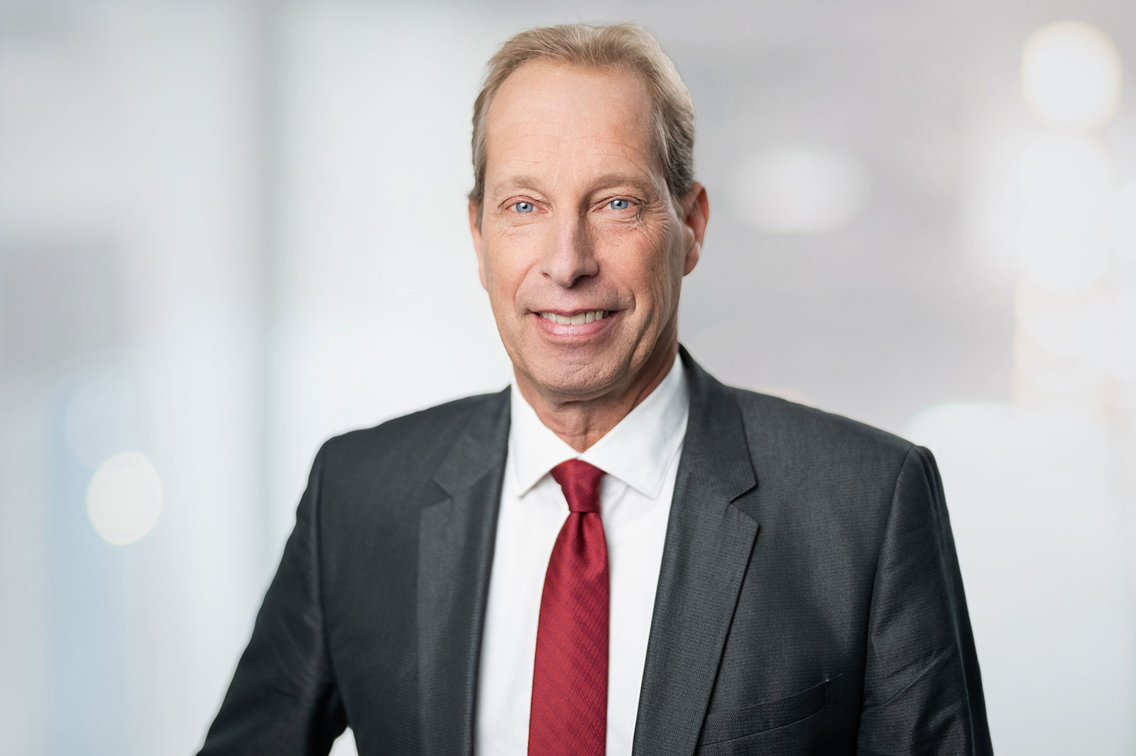 A man in a suit with a red tie stands against a blurred background.