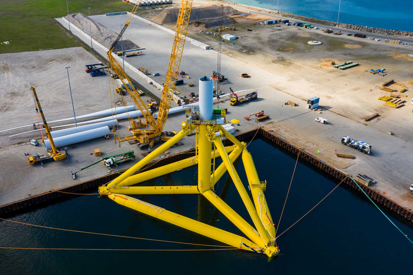 Ein gelbes Fundament eines Windkraftwerks im Wasser, umgeben von Baugeräten und Rohren auf dem Land.