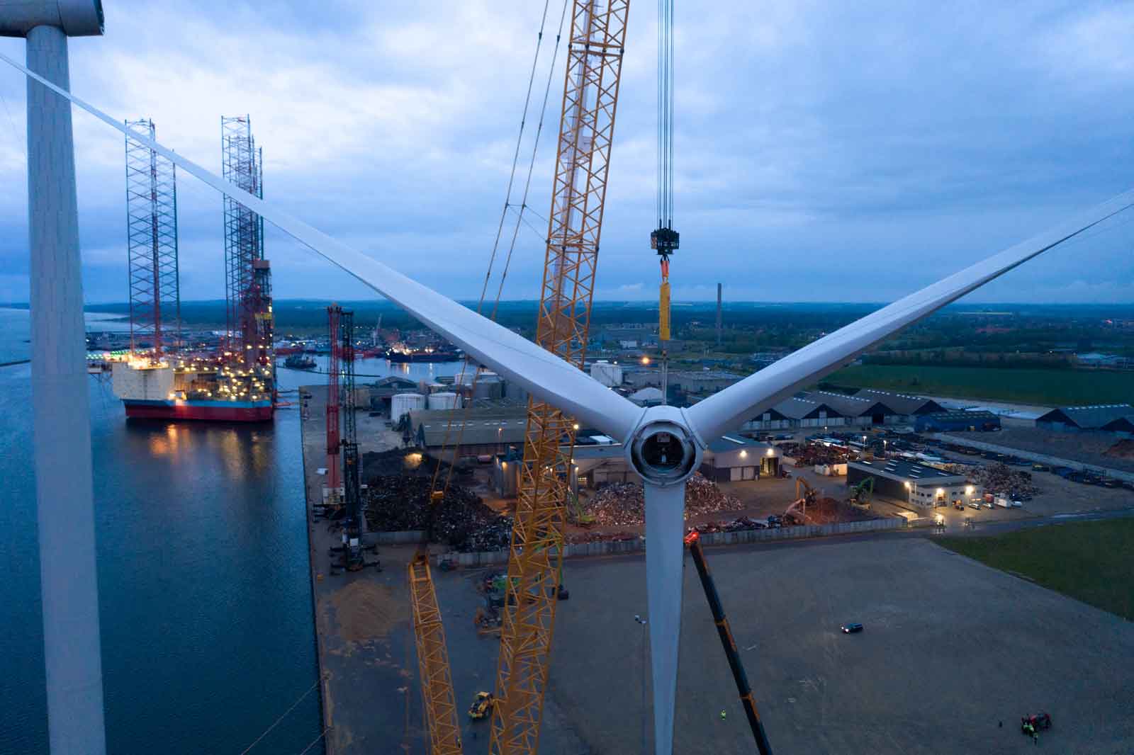 Blick auf einen Windkraftanlagenbauplatz mit Kränen und Schiffen im Hafen unter bewölktem Himmel.