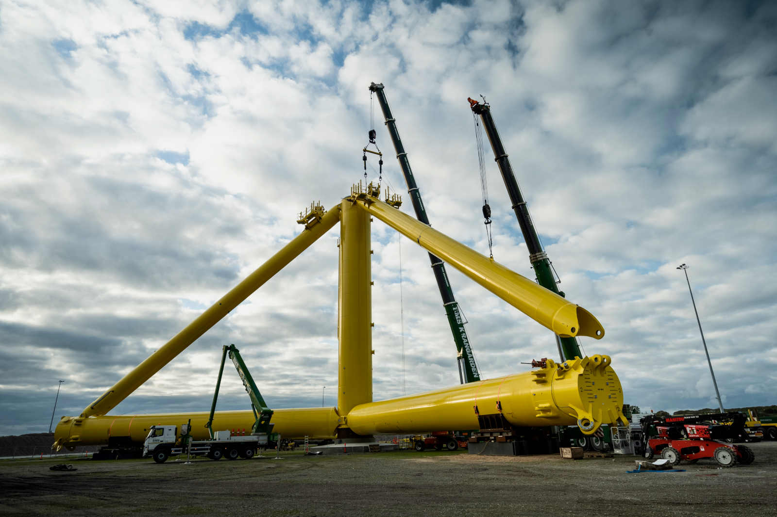 Große gelbe Konstruktionsteile werden von Kränen auf einer Baustelle angehoben, mit einem bewölkten Himmel im Hintergrund.