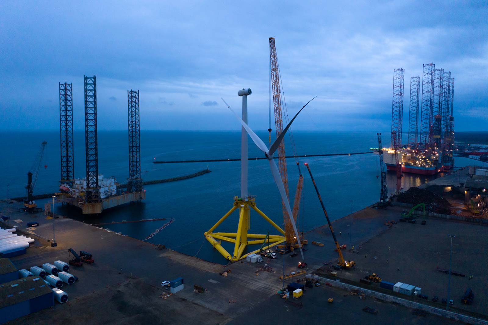 Ein Offshore-Windpark mit einem großen Windrad, Kräne und ein Ozean im Hintergrund bei Dämmerung.
