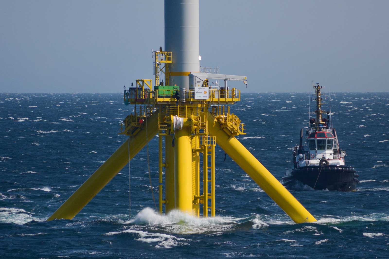 Ein gelbes Offshore-Windkraftwerk steht im Meer, während ein Schlepper in der Nähe fährt.