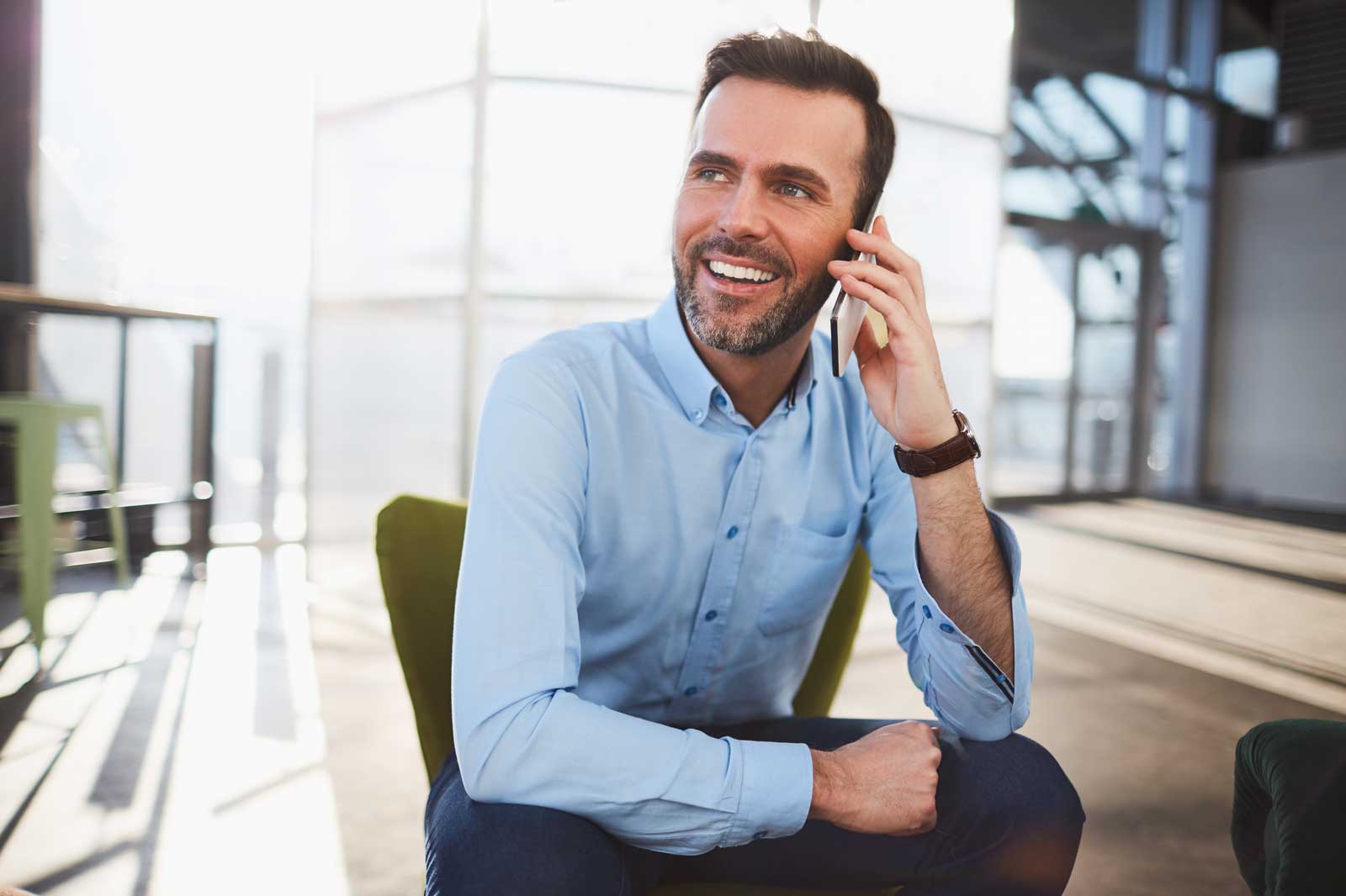 A man in a light blue shirt sits in a chair, talking on the phone in a modern office setting.