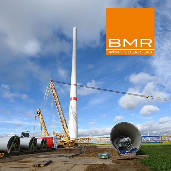 A wind turbine is being constructed, featuring cranes and turbine components in the foreground under a cloudy sky.